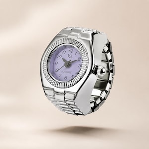 Puede incluir: Un anillo reloj de tono plateado con una esfera redonda de color morado claro, números negros, las letras Ks y la palabra QUARTZ. La banda está diseñada para parecerse a una correa de reloj de eslabones metálicos con un bisel texturizado.