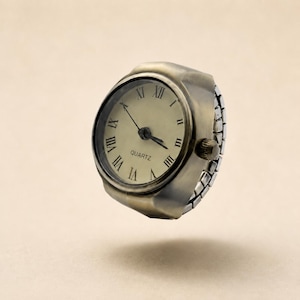 Può includere: Un anello orologio con cassa rotonda in metallo color ottone antico e quadrante color crema. Il quadrante presenta numeri romani neri, lancette nere sottili e la scritta QUARTZ stampata in nero. È dotato di un cinturino flessibile color argento.