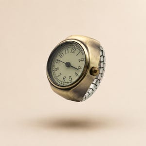 Peut inclure: Une bague montre dotée d'un boîtier rond en métal couleur laiton antique et d'un cadran crème. Le cadran affiche des chiffres arabes noirs et deux fines aiguilles noires. Elle est munie d'un bracelet extensible en métal argenté.