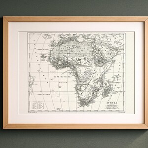 Peut inclure: Une carte ancienne de l'Afrique encadrée, imprimée en noir et blanc sur un fond crème. La carte présente des détails géographiques précis, des lignes de latitude et de longitude, ainsi que le texte AFRIKA en bas à droite, dans un cadre en bois clair.