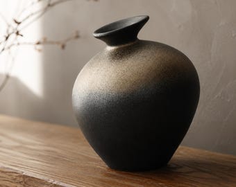 Pode incluir: Um vaso de cerâmica redondo com pescoço estreito e borda alargada. O vaso apresenta um acabamento texturizado que transita de um preto fosco profundo na base para um gradiente dourado metálico na parte superior, criando um visual bicolor.