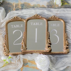 May include: Three arched table number signs with ornate gold-toned frames. Each mirror features the word TABLE printed in white above the numbers 2, 1, and 3 respectively. The frames have a rope-like border and decorative scrollwork at the top.