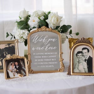 May include: A gold ornate arched frame containing a memorial sign with white text that reads: Wish you were here, IN LOVING MEMORY OF THOSE WHO CANNOT BE HERE TODAY BUT ARE FOREVER PRESENT IN OUR HEARTS. Two additional gold frames hold vintage photos.