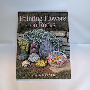 May include: A softcover book titled Painting Flowers on Rocks by Lin Wellford. The cover features a collection of stones painted with various floral designs, including yellow daffodils, purple tulips, and a basket arrangement of colorful flowers.