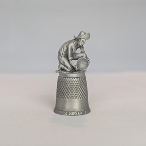 Peut inclure: Un dé à coudre en métal argenté surmonté d'une figurine sculptée représentant une personne portant un chapeau à larges bords, agenouillée et tenant une bassine circulaire. La base présente un motif alvéolé et une bande décorative.