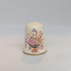 May include: A cream-colored ceramic thimble featuring a colorful floral arrangement in a basket design. The pattern includes pink, orange, and blue flowers with green stems. The base of the thimble is finished with a thin, metallic gold rim.