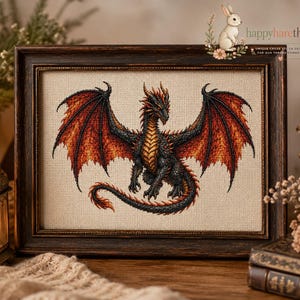 May include: A framed cross-stitch embroidery featuring a central dragon with black scales, a golden underbelly, and large wings transitioning from black to vibrant orange and red, set against a light beige fabric background within a dark wood frame.