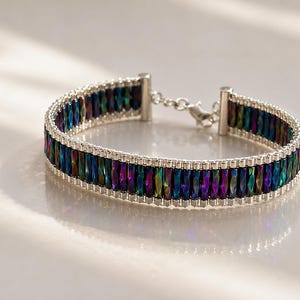 Könnte beinhalten: Ein Armband mit einer Reihe schillernder, mehrfarbiger rechteckiger Perlen mit metallischem Finish. Die Perlen sind von kleinen silberfarbenen Saatperlen umrandet und durch silberfarbene Metallendstücke mit einem Karabinerverschluss verbunden.