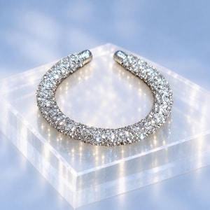 Puede incluir: Un brazalete abierto de tono plateado con una superficie continua cubierta de pequeños cristales claros y reflectantes. La banda circular tiene una pequeña abertura en la parte superior, creando una forma similar a una herradura.