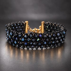 Könnte beinhalten: Ein breites Armband aus mehreren Reihen facettierter schwarzer Perlen mit irisierenden blauen und violetten Reflexen. Das Schmuckstück verfügt über einen goldfarbenen Metallverschluss in der Mitte, der die beiden Enden des Perlenbandes sicher verbindet.