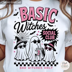 May include: A white t-shirt featuring a graphic with the text BASIC Witches SOCIAL CLUB and BAD WITCHES ONLY. The design includes three ghosts wearing witch hats and heart-shaped sunglasses, a spider web, bats, stars, and a black and white checkerboard.