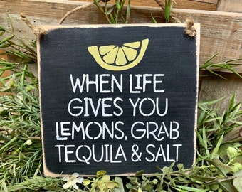 May include: A black wooden sign with white text that reads "When life gives you lemons, grab tequila & salt". A yellow lemon slice graphic is above the text.
