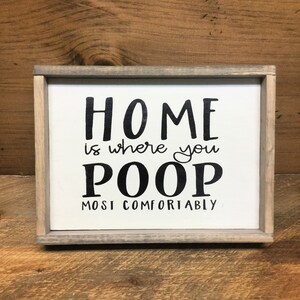 Puede incluir: Un letrero enmarcado con el texto "HOME is where you POOP most comfortably" en letras negras sobre un fondo blanco. El marco es de color gris claro.