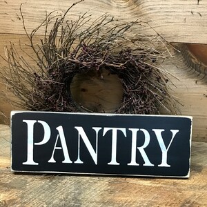 May include: Black wooden sign with white lettering that reads "PANTRY". The sign is sitting on a wooden surface with a brown twig wreath in the background.