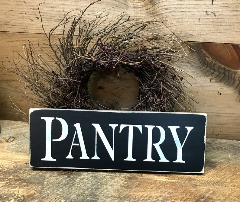 May include: Black wooden sign with white lettering that reads "PANTRY". The sign is sitting on a wooden surface with a brown twig wreath in the background.
