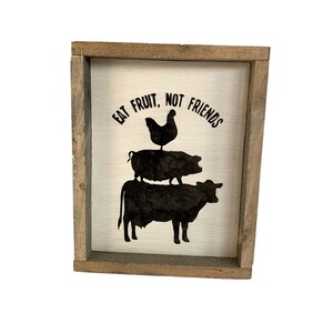 May include: A white wooden sign with a brown frame, featuring a black silhouette of a cow, pig, and chicken stacked on top of each other. The sign reads "Eat Fruit, Not Friends".