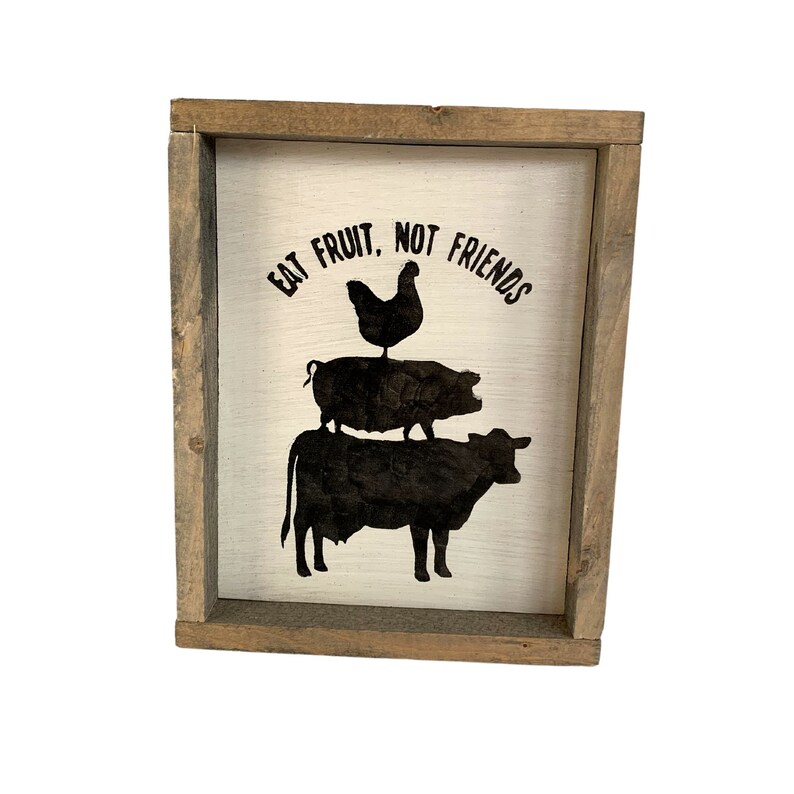 May include: A white wooden sign with a brown frame, featuring a black silhouette of a cow, pig, and chicken stacked on top of each other. The sign reads "Eat Fruit, Not Friends".