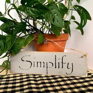 May include: A gray wooden sign with the word "Simplify" written in black cursive lettering. The sign is hanging from a twine string and is in front of a potted plant with green leaves.