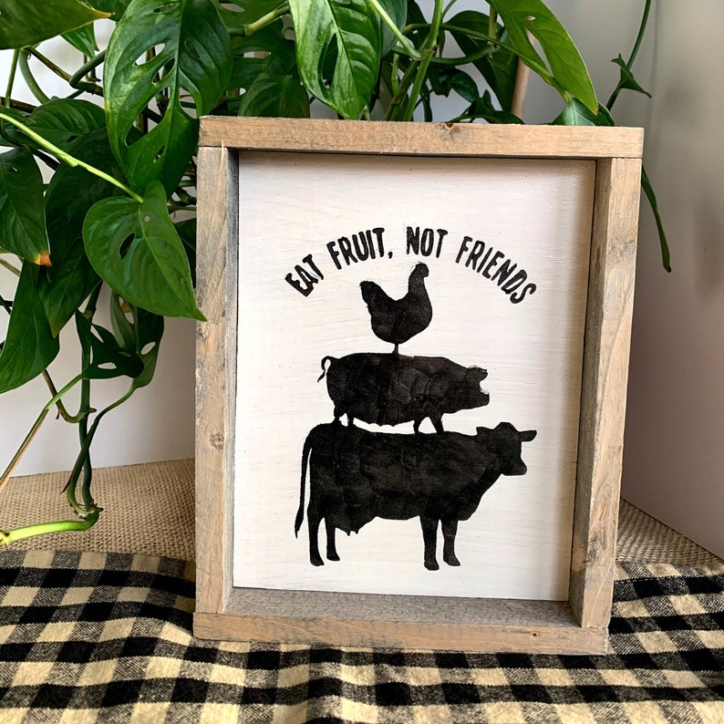 May include: A white wooden sign with black silhouettes of a cow, pig, and chicken stacked on top of each other. The sign reads "Eat Fruit, Not Friends".