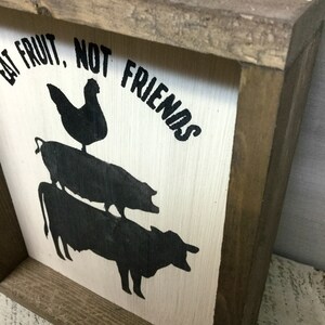 May include: A white wooden sign with black painted silhouettes of a chicken, pig, and cow stacked on top of each other. The sign reads "Eat Fruit, Not Friends".