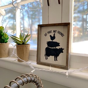 May include: A white wooden sign with black text and silhouettes of a cow, pig, and chicken. The sign reads "Eat Fruit, Not Friends".