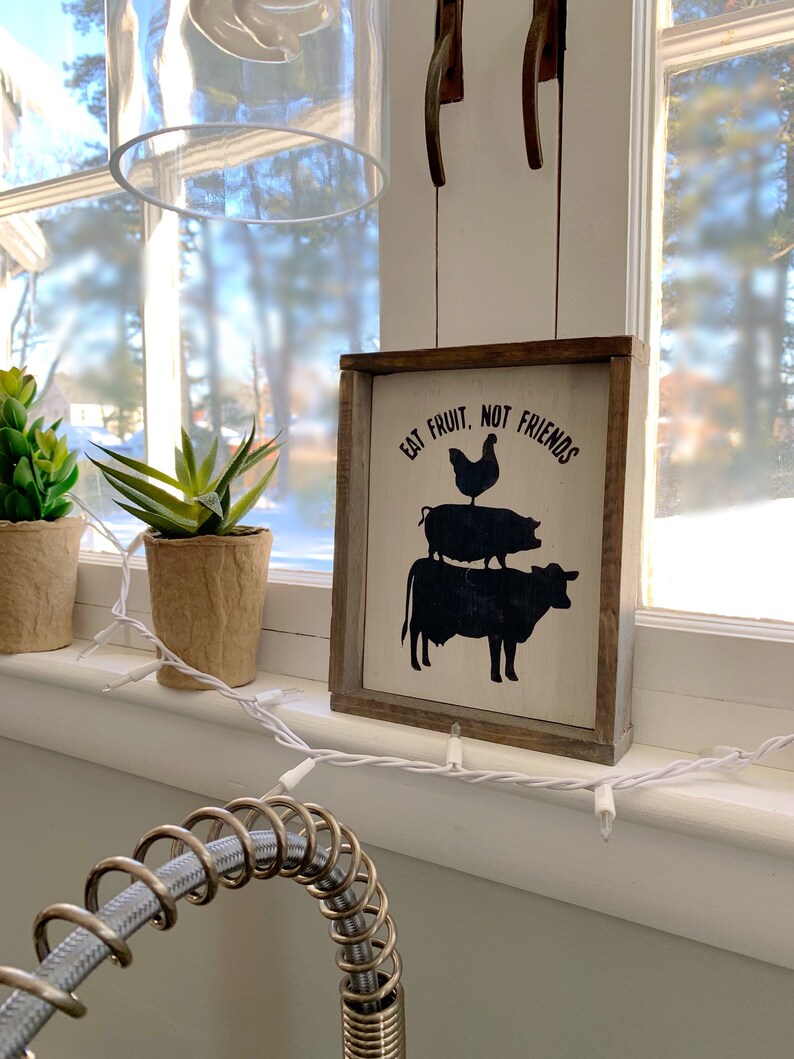 May include: A white wooden sign with black text and silhouettes of a cow, pig, and chicken. The sign reads "Eat Fruit, Not Friends".