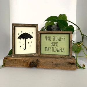 May include: Two wooden signs with a rustic brown finish. The sign on the left has a black silhouette of an umbrella with raindrops. The sign on the right has a green background with the text "April showers bring May flowers".