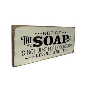 May include: A white wooden sign with black lettering that reads "...NOTICE... THE SOAP IS NOT JUST FOR DECORATION PLEASE USE IT! - "
