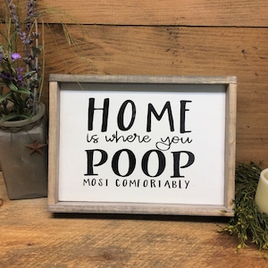 Puede incluir: Un letrero enmarcado con el texto "HOME is where you POOP MOST COMFORTABLY" en letras negras. El letrero tiene un fondo blanco y un marco de madera gris. Elementos decorativos incluyen un jarr&oacute;n con flores moradas.