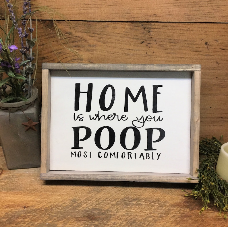 Puede incluir: Un letrero enmarcado con el texto "HOME is where you POOP MOST COMFORTABLY" en letras negras. El letrero tiene un fondo blanco y un marco de madera gris. Elementos decorativos incluyen un jarr&oacute;n con flores moradas.