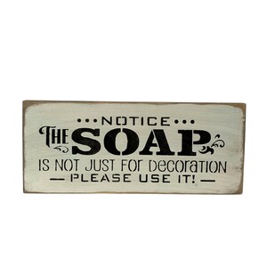 May include: A white wooden sign with black text that reads "NOTICE... THE SOAP IS NOT JUST FOR DECORATION - PLEASE USE IT! - "