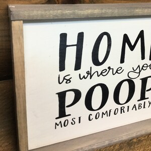 Puede incluir: Un letrero rectangular de madera con fondo blanco y marco gris. El letrero presenta texto escrito a mano en negro que dice "HOME is where you POOP most comfortably".