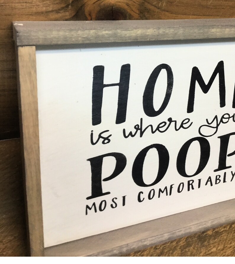 Puede incluir: Un letrero rectangular de madera con fondo blanco y marco gris. El letrero presenta texto escrito a mano en negro que dice "HOME is where you POOP most comfortably".