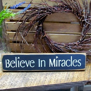 May include: A black wooden sign with white lettering that reads "Believe In Miracles". The sign is sitting on a wooden surface with a twig wreath behind it.