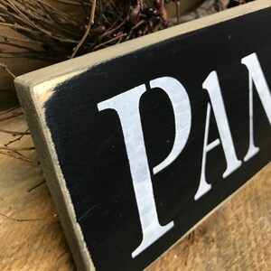 May include: A black wooden sign with white painted letters spelling "PANTRY". The sign has a distressed, rustic look.