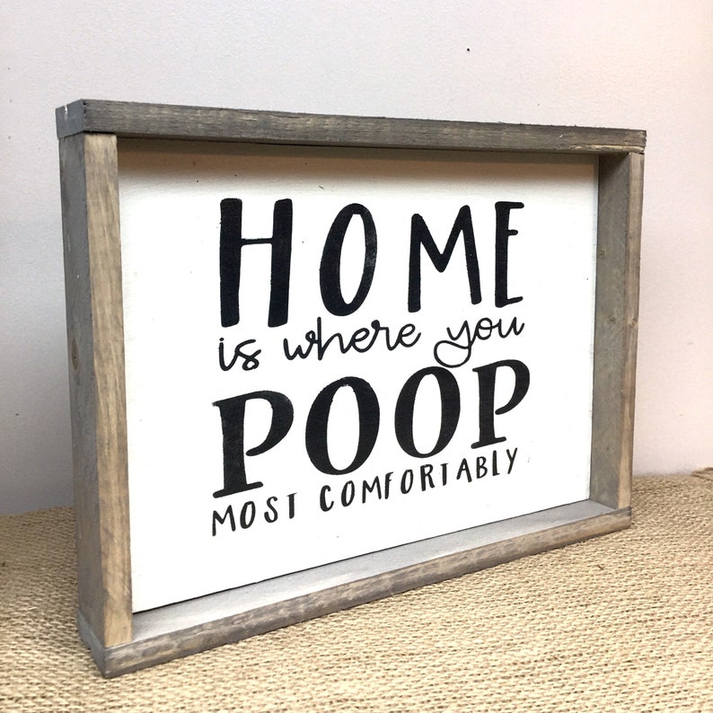 Puede incluir: Un letrero de madera enmarcado con el texto "HOME is where you POOP most comfortably" en letras negras sobre un fondo blanco. El marco es de color gris desgastado.