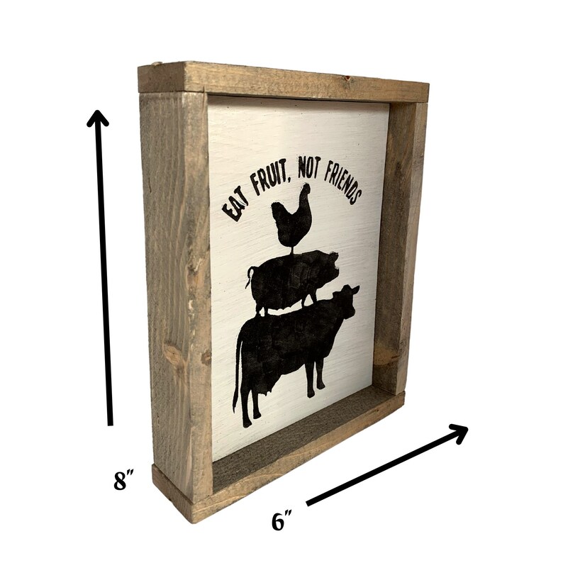 May include: A white wooden framed sign with the text "Eat Fruit, Not Friends" in black lettering. The sign features a black silhouette of a chicken standing on a pig standing on a cow. The sign measures 6 inches wide by 8 inches tall.
