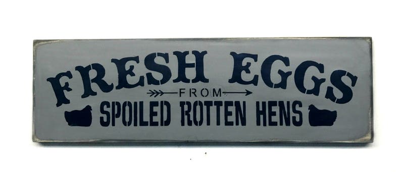 May include: A gray wooden sign with black text that reads "FRESH EGGS FROM SPOILED ROTTEN HENS". The sign has a distressed look and features two stylized chicken silhouettes.