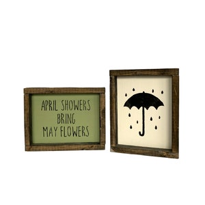 May include: Two wooden framed signs with a spring theme. The first sign has a green background with the text "APRIL SHOWERS BRING MAY FLOWERS". The second sign has a white background with a black silhouette of an umbrella with raindrops.