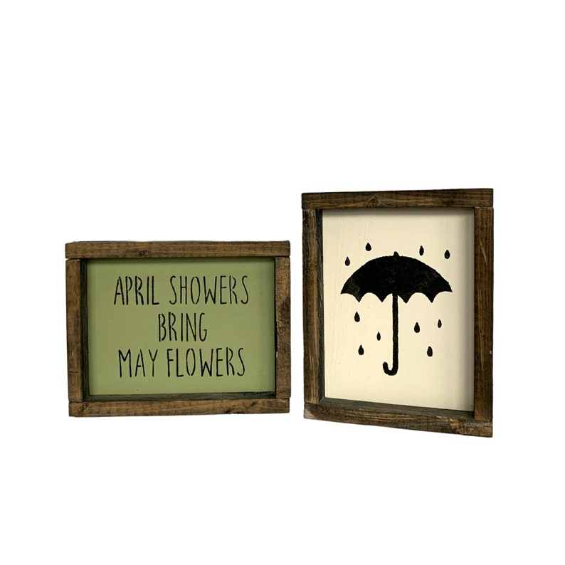 May include: Two wooden framed signs with a spring theme. The first sign has a green background with the text "APRIL SHOWERS BRING MAY FLOWERS". The second sign has a white background with a black silhouette of an umbrella with raindrops.