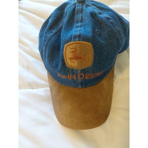 May include: A blue denim baseball cap featuring a tan patch with a brown leaping deer logo and the text JOHN DEERE embroidered in brown thread. The cap has a solid tan suede-like brim and a structured crown with a single ventilation eyelet.