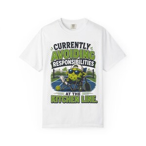 May include: A white t-shirt featuring a graphic of a pickleball character wearing sunglasses and a cap, sitting in a folding chair on a court. The text reads: CURRENTLY AVOIDING RESPONSIBILITIES AT THE KITCHEN LINE. A pickleball paddle and ball are nearby.