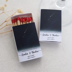 May include: Two rectangular matchboxes featuring a dark night sky design with a shooting star. The text reads WHERE OUR STORY BEGAN, Amelia & Theodore, JULY 10, 2027, and coordinates. One box is open, revealing wooden matches with red tips.