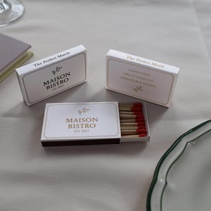 May include: Three white rectangular matchboxes with gold borders and text. Two boxes are closed, displaying "The Perfect Match" and "MAISON BISTRO EST. 2027". One open box reveals wooden matches with red tips, featuring the same branding and a small leaf icon.