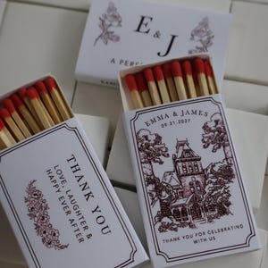 May include: Two white matchboxes with red-tipped matches. One box features a maroon illustration of a house, the text "EMMA & JAMES 09.21.2027" and "THANK YOU FOR CELEBRATING WITH US". The other box displays "THANK YOU LOVE, LAUGHTER & HAPPY EVER AFTER".