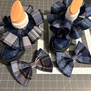 May include: Two navy blue patent leather boots featuring ruffled blue, grey, and yellow plaid fabric cuffs around the ankles. Two matching plaid fabric bow accessories are positioned in front of the boots, each featuring the same blue, grey, and yellow pattern.
