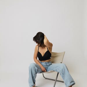 Peut inclure: Une brassière courte noire avec de fines bretelles et un petit logo blanc sur le côté, associée à un jean bleu clair en denim à coupe large avec une fermeture classique à boutons et à glissière, portée en étant assis sur une chaise pliante beige.
