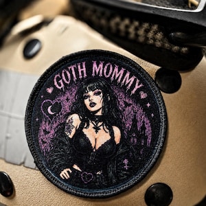 May include: A circular embroidered patch featuring a dark-haired figure in black clothing with the text GOTH MOMMY arched above. The design includes purple accents, a heart, a crescent moon, and a silhouette of a building against a dark background.