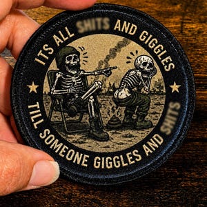 May include: A circular embroidered patch with a black border featuring two skeletons in a desert setting. One skeleton sits in a chair pointing, while the other squats. The text reads: ITS ALL SHITS AND GIGGLES TILL SOMEONE GIGGLES AND SHITS.