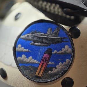 May include: A circular embroidered patch featuring a blue sky background with white clouds and a dark grey fighter jet flying above a gradient pink and orange vape device that has a small smiley face icon and the word HAPPINESS printed on it.
